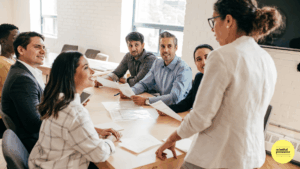 6 people sitting around a table in meeting room with one woman standing