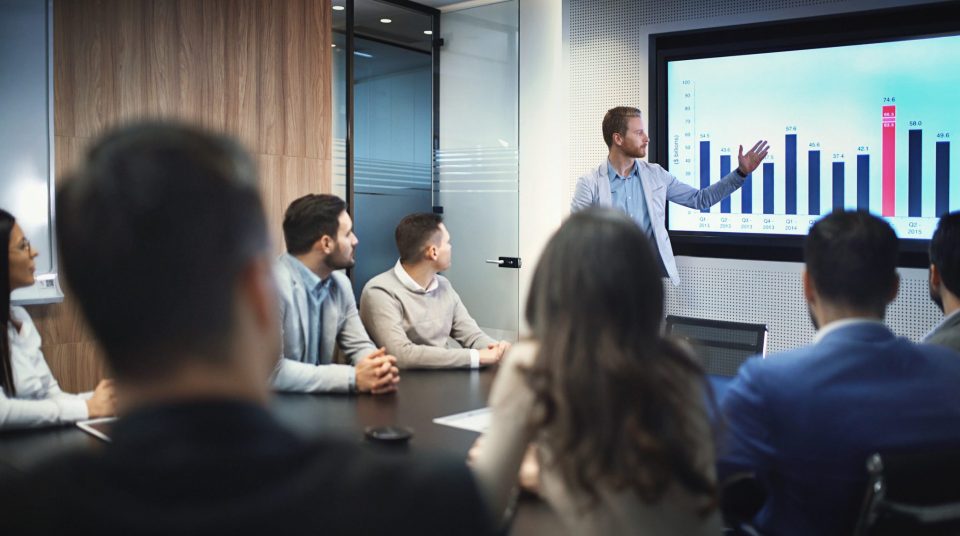 Closeup of group of business people in a meeting seated around squared office desk. Several people are seated around conference table and team leader is having a presentation in front of a projection screen. He's showing annual profit charts ending with 2016 result.