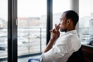 Pensive and serious young black businessman sitting in a chair in his office and looking through a window away. Shot from behind.