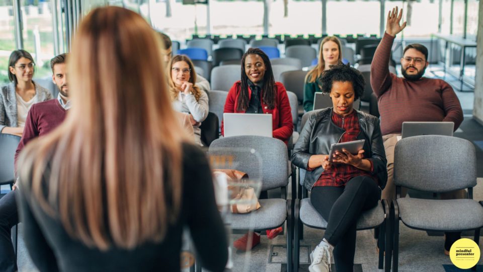 woman presenting to an audience and one man in the audience has his hand raised.
