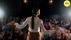 woman standing on a stage facing and audience with here arms wide open