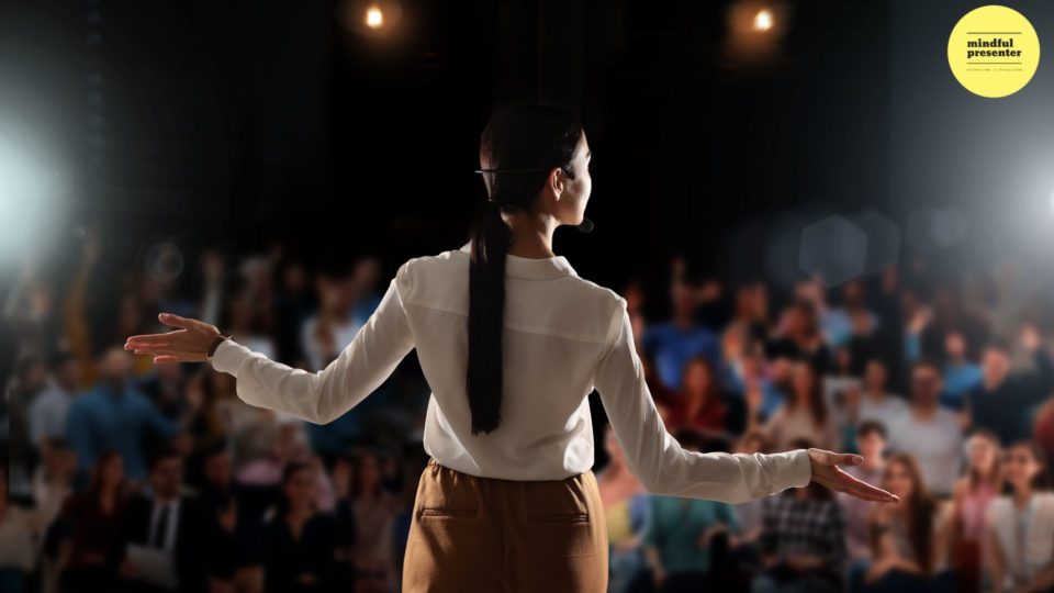 woman standing on a stage facing and audience with here arms wide open