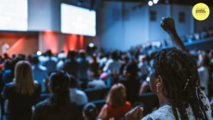 blurred audience with screen at front of stage and a woman raising her hand at the back